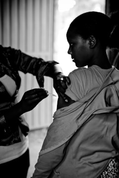 A person is administering a vaccination shot to a young woman. The young woman has her shirt sleeve rolled up, and the scene is captured in black and white, highlighting the somber and serious nature of the moment.
