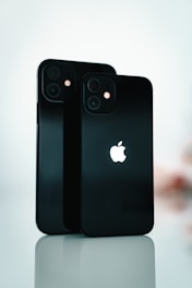 Two black smartphones are standing upright on a reflective surface. The Apple logo is prominently displayed on both devices. The background is softly blurred, emphasizing the sleek design and the glossy finish of the phones.