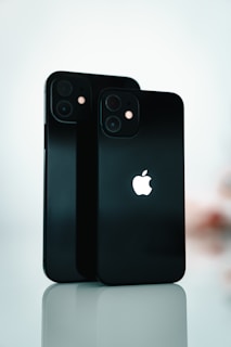 Two black smartphones are standing upright on a reflective surface. The Apple logo is prominently displayed on both devices. The background is softly blurred, emphasizing the sleek design and the glossy finish of the phones.