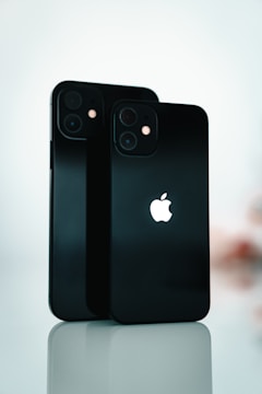 Two black smartphones are standing upright on a reflective surface. The Apple logo is prominently displayed on both devices. The background is softly blurred, emphasizing the sleek design and the glossy finish of the phones.