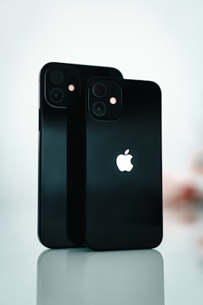 Two black smartphones are standing upright on a reflective surface. The Apple logo is prominently displayed on both devices. The background is softly blurred, emphasizing the sleek design and the glossy finish of the phones.