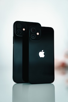 Two black smartphones are standing upright on a reflective surface. The Apple logo is prominently displayed on both devices. The background is softly blurred, emphasizing the sleek design and the glossy finish of the phones.