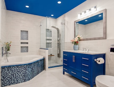 A modern bathroom features a large glass-enclosed shower with metallic fixtures and a bathtub adorned with mosaic tiles. A vibrant blue vanity with silver handles stands against the wall, topped with a marble counter and a large mirror. Bright lights illuminate the space, and a bouquet of flowers in a blue vase adds a touch of color.