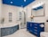 A modern bathroom features a large glass-enclosed shower with metallic fixtures and a bathtub adorned with mosaic tiles. A vibrant blue vanity with silver handles stands against the wall, topped with a marble counter and a large mirror. Bright lights illuminate the space, and a bouquet of flowers in a blue vase adds a touch of color.