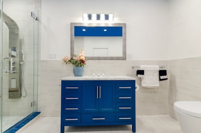 A modern bathroom featuring a blue vanity with a marble countertop and a large framed mirror above it. On the countertop, there's a blue vase holding pink and white flowers. To the left, a glass-enclosed shower with chrome fixtures is visible. A toilet sits to the right with a double towel rack on the wall holding white and black towels.