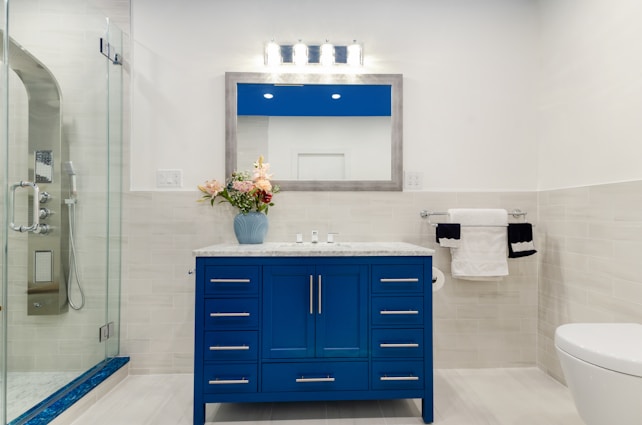 A modern bathroom featuring a blue vanity with a marble countertop and a large framed mirror above it. On the countertop, there's a blue vase holding pink and white flowers. To the left, a glass-enclosed shower with chrome fixtures is visible. A toilet sits to the right with a double towel rack on the wall holding white and black towels.