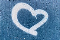 A white heart shape is spray-painted on a textured blue wall. The heart appears to have a slight drip effect at the top, suggesting a graffiti art style.