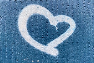 A white heart shape is spray-painted on a textured blue wall. The heart appears to have a slight drip effect at the top, suggesting a graffiti art style.
