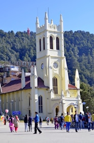 A historic yellow church with Gothic-style architecture is situated in an open area. Surrounding the church are several people walking and engaging in activities. In the background, there is dense greenery with tall trees and a clear blue sky. Some colorful shops and a statue can also be seen amidst the greenery.