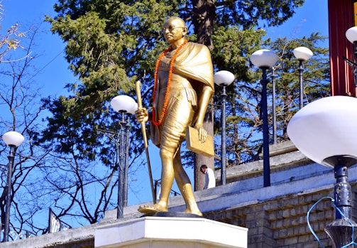 A golden statue of a man dressed in traditional clothing stands on a pedestal. The statue is adorned with an orange garland and holds a book in one hand and a walking stick in the other. The statue is set against a backdrop of tall trees and various lamp posts.