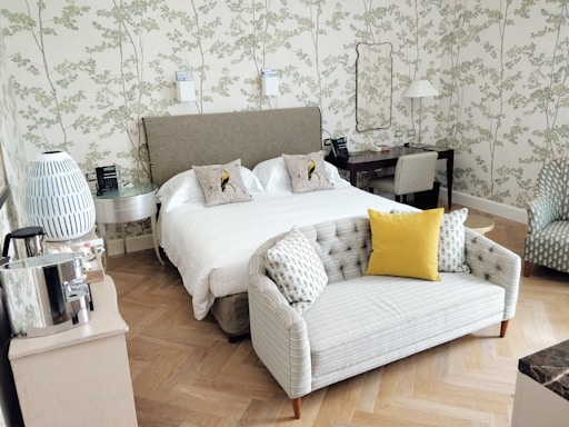 A well-furnished bedroom featuring a double bed with white linens and two decorative pillows on a soft headboard. A small, gray sofa with a bright yellow pillow sits at the foot of the bed. The walls are adorned with a subtle floral pattern. There is a lamp and telephone on the bedside table, and a small desk in the corner with a chair. A decorative lamp with a blue and white pattern and a silver coffee maker are on a side table.