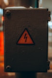 A weathered electrical box with a yellow warning sign featuring a lightning bolt symbol, indicating caution. The box is metallic and appears to be placed outside, showing signs of wear.