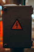 A weathered electrical box with a yellow warning sign featuring a lightning bolt symbol, indicating caution. The box is metallic and appears to be placed outside, showing signs of wear.