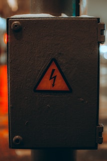 A weathered electrical box with a yellow warning sign featuring a lightning bolt symbol, indicating caution. The box is metallic and appears to be placed outside, showing signs of wear.