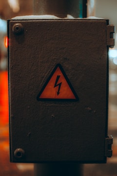 A weathered electrical box with a yellow warning sign featuring a lightning bolt symbol, indicating caution. The box is metallic and appears to be placed outside, showing signs of wear.