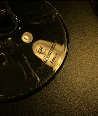 A close-up view of a wine glass base with the words 'Chef & Sommelier France' printed on it in elegant, dark font. The glass is placed on a textured, dark surface, reflecting a warm light.