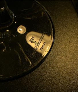 A close-up view of a wine glass base with the words 'Chef & Sommelier France' printed on it in elegant, dark font. The glass is placed on a textured, dark surface, reflecting a warm light.