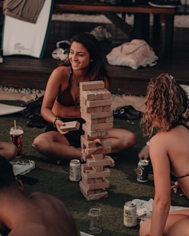 A group of people are sitting on the ground outdoors, engaged in a game of Jenga with a tall stack of wooden blocks. One person is holding a block, smiling and interacting with others around them. Various items like a surfboard, drinks, and bags are scattered in the background, indicating a relaxed and casual setting.