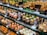 A display of various tapas arranged neatly on trays in a food counter. The tapas include a variety of ingredients such as quail eggs, burgers, goat cheese, vegetables, and assorted seafood, all skewered and labeled with descriptions in both Spanish and English. The vibrant and colorful selection is presented on a multi-tiered glass shelf, showcasing a rich assortment of textures and flavors.