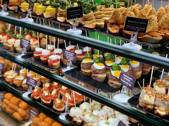 A display of various tapas arranged neatly on trays in a food counter. The tapas include a variety of ingredients such as quail eggs, burgers, goat cheese, vegetables, and assorted seafood, all skewered and labeled with descriptions in both Spanish and English. The vibrant and colorful selection is presented on a multi-tiered glass shelf, showcasing a rich assortment of textures and flavors.