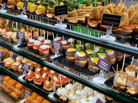 A display of various tapas arranged neatly on trays in a food counter. The tapas include a variety of ingredients such as quail eggs, burgers, goat cheese, vegetables, and assorted seafood, all skewered and labeled with descriptions in both Spanish and English. The vibrant and colorful selection is presented on a multi-tiered glass shelf, showcasing a rich assortment of textures and flavors.