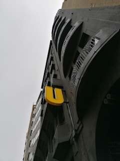 A tall building with a modern architectural style, featuring multiple balconies with metal railings. A prominent, large yellow letter 'U' is attached to the building's facade, creating a striking contrast against the dark, industrial design of the structure. The sky is overcast, emphasizing the building's imposing height and presence.