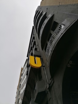 A tall building with a modern architectural style, featuring multiple balconies with metal railings. A prominent, large yellow letter 'U' is attached to the building's facade, creating a striking contrast against the dark, industrial design of the structure. The sky is overcast, emphasizing the building's imposing height and presence.