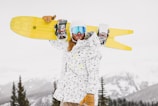 A person is standing outdoors in a snowy mountainous area, wearing a white and black speckled jacket, brown gloves, and reflective blue goggles. They are holding a yellow snowboard over their shoulder, and their long hair is visible. Pine trees and snow-covered peaks are in the background.