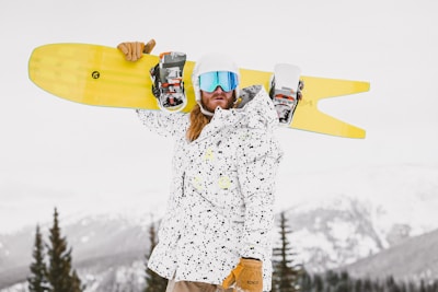 A person is standing outdoors in a snowy mountainous area, wearing a white and black speckled jacket, brown gloves, and reflective blue goggles. They are holding a yellow snowboard over their shoulder, and their long hair is visible. Pine trees and snow-covered peaks are in the background.