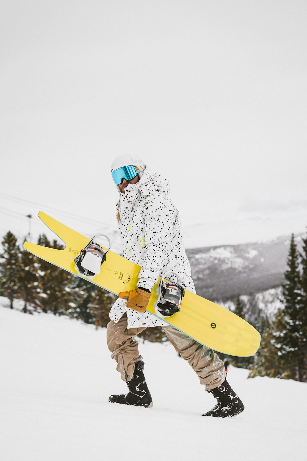 Mann in weiß-grüner Jacke und brauner Hose, der tagsüber ein gelbes Snowboard hält