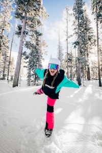 A person dressed in colorful winter sports attire, including a helmet and reflective goggles, playfully striking a pose on a snowy landscape surrounded by tall, snow-covered pine trees.