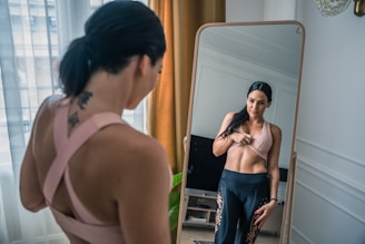 A woman with a tattoo on her back is looking at herself in a full-length mirror. She is wearing a pink sports bra and dark leggings with decorative patterns. The setting is a well-lit room with a window covered by sheer curtains and a mustard-colored drape. The woman is adjusting her sports bra while smiling subtly at her reflection.