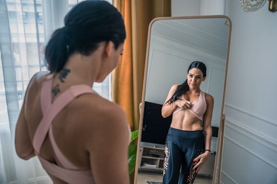 A woman with a tattoo on her back is looking at herself in a full-length mirror. She is wearing a pink sports bra and dark leggings with decorative patterns. The setting is a well-lit room with a window covered by sheer curtains and a mustard-colored drape. The woman is adjusting her sports bra while smiling subtly at her reflection.
