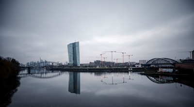 A modern cityscape with a prominent skyscraper adjacent to a river, surrounded by several construction cranes. The overcast sky and the buildings reflect on the calm water surface, while a bridge spans the river. The image captures an urban development scene with cloudy weather conditions.