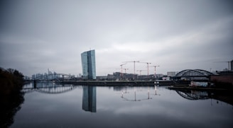 A modern cityscape with a prominent skyscraper adjacent to a river, surrounded by several construction cranes. The overcast sky and the buildings reflect on the calm water surface, while a bridge spans the river. The image captures an urban development scene with cloudy weather conditions.