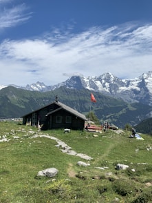 brown wooden house on green grass field near mountain under white clouds and blue sky during