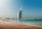 A modern, sail-shaped skyscraper stands prominently by the beachside, with people enjoying the seaside in the foreground. A clear blue sky and turquoise waters complement the sandy beach, creating a picturesque coastal scene.
