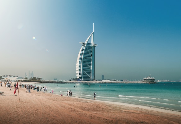 A modern, sail-shaped skyscraper stands prominently by the beachside, with people enjoying the seaside in the foreground. A clear blue sky and turquoise waters complement the sandy beach, creating a picturesque coastal scene.
