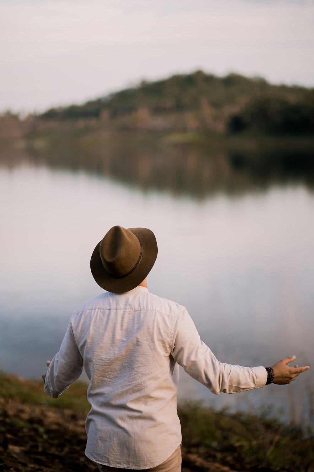 person in white long sleeve shirt wearing brown fedora hat standing near lake during daytime