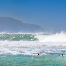 Several surfers are riding large, powerful waves in an expansive ocean setting. The sky is clear and blue, with distant mountains visible on the horizon. The water is a vibrant mix of green and blue, with white foam cresting on the waves.