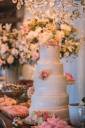 A beautifully decorated four-tier wedding cake adorned with intricate lace patterns and pink floral arrangements on each tier. The cake is situated on a richly decorated table with an assortment of appetizers and decorative pink rose petals. Above, a luxurious chandelier with crystal pendants adds a touch of elegance, while a background of white and pink flowers complements the festive setting.