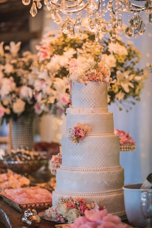 A beautifully decorated four-tier wedding cake adorned with intricate lace patterns and pink floral arrangements on each tier. The cake is situated on a richly decorated table with an assortment of appetizers and decorative pink rose petals. Above, a luxurious chandelier with crystal pendants adds a touch of elegance, while a background of white and pink flowers complements the festive setting.