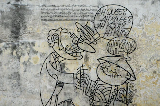 A mural featuring a cartoonish metal wire sculpture on a textured, weathered wall. Two figures are depicted: one with a mustache and a suit, the other wearing a traditional hat and beaded necklace. Speech bubbles contain text, contributing to the humorous tone.