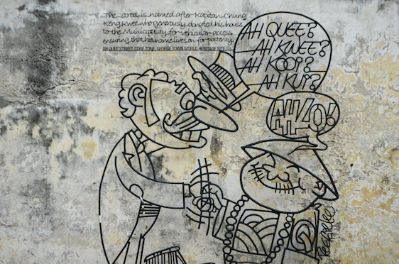 A mural featuring a cartoonish metal wire sculpture on a textured, weathered wall. Two figures are depicted: one with a mustache and a suit, the other wearing a traditional hat and beaded necklace. Speech bubbles contain text, contributing to the humorous tone.