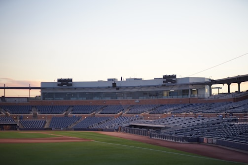 An empty baseball stadium with rows of blue seats surrounding the field. The stadium has a large central building with windows and a scoreboard on top. The sky is a soft, gradient of light colors as the sun sets.