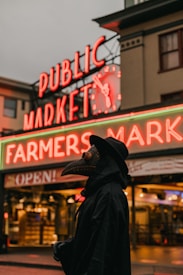 A person dressed in a dark plague doctor outfit stands in front of a brightly lit sign reading 'Public Market' and 'Farmers Market' at night. The background features a neon clock and storefronts, enhancing a mysterious, historical atmosphere.
