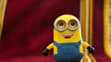 A yellow character with goggles and a blue outfit stands with a cheerful expression against a deep red background with gold accents.