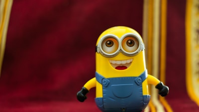 A yellow character with goggles and a blue outfit stands with a cheerful expression against a deep red background with gold accents.