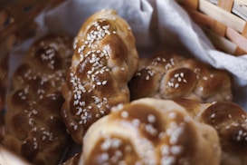 Freshly baked braided bread rolls sprinkled with sesame seeds are arranged in a wicker basket lined with cloth. The bread has a golden-brown crust, indicating a perfect bake.
