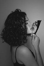 A black and white portrait features a person with curly hair holding a broken mirror shard. The reflection in the shard shows part of their face, creating a dramatic and introspective effect.